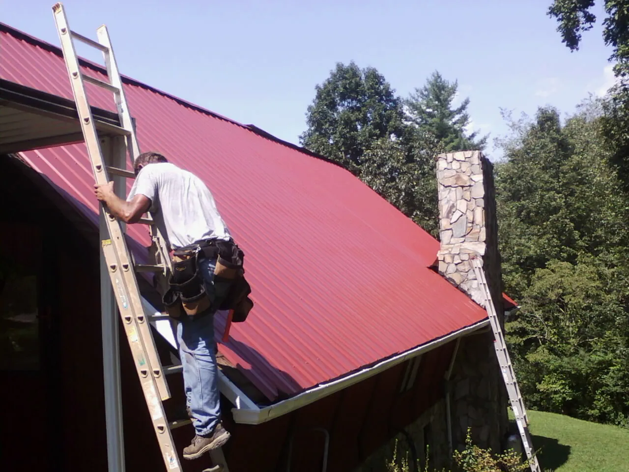 Roofer on a ladder beside a red metal roof and stone chimney — Soffit & Fascia Repair by Roofing Brothers in West Chicago